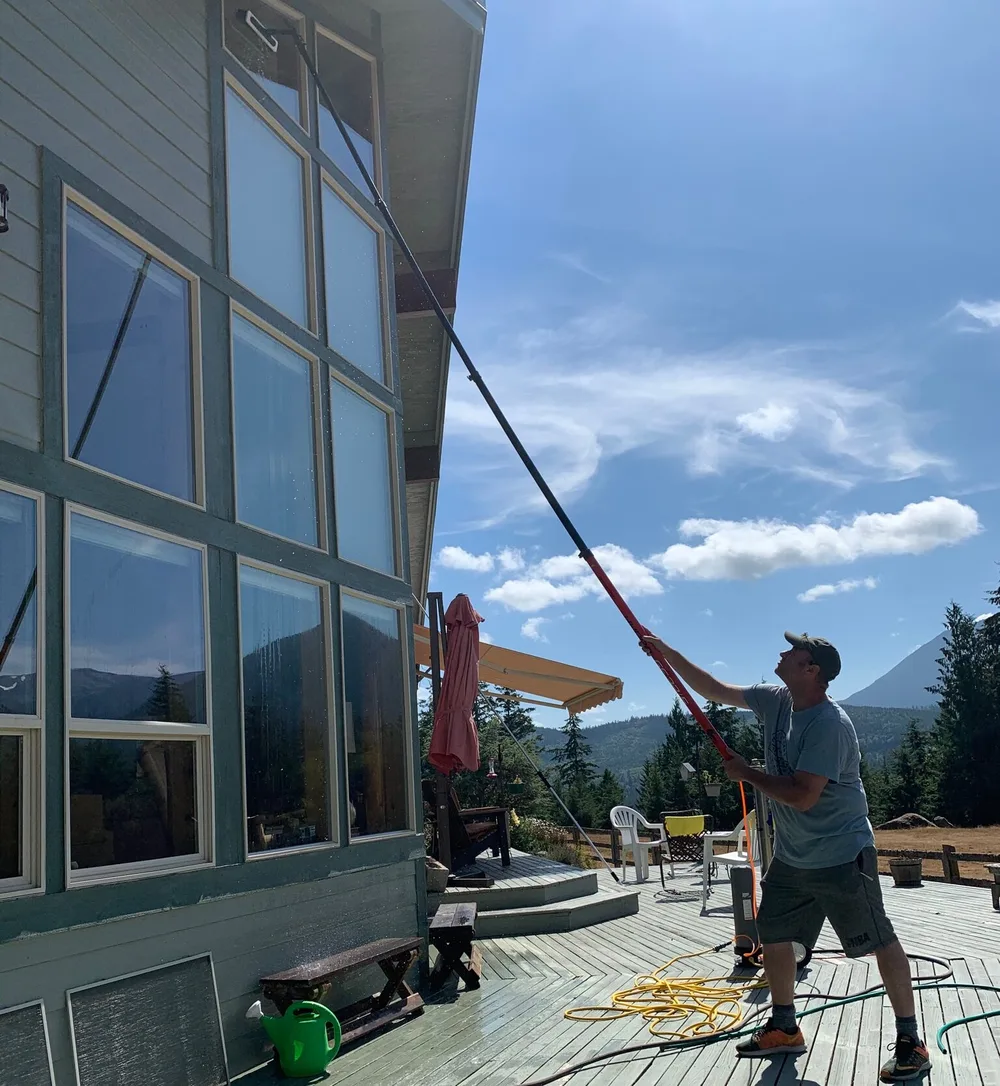 Cleaning tall windows on a house with a water-fed pole from the deck, mountains in the distance